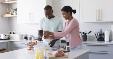 Çeşitli bir çift modern bir mutfakta birlikte kahvaltı hazırlıyor. Afrika kökenli Amerikalı koca ve eşi, ikisi de genç yetişkinler, taze malzemelerle çevrili, değişmemiş yiyeceklere odaklanmışlar.