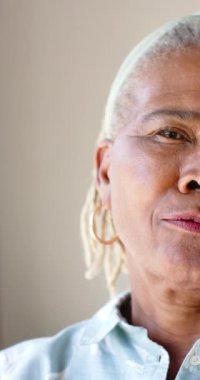 Dikey video: Yaşlı bir Afrikalı Amerikalı kadın gülümsemeyle yan yana bakıyor. Kısa, beyaz rastaları, altın halka küpeleri ve açık mavi bir gömleği var.