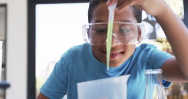 Bir okul fen laboratuvarı sınıfında, genç bir Afrikalı Amerikalı öğrenci bir deney yapıyor. Güvenlik gözlüğü takıyor, mavi bir gömlek giyiyor ve değiştirilmemiş bir şişeye sıvı dökmeye odaklanıyor..