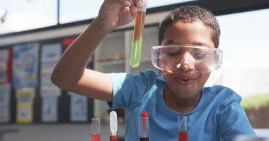 Okulda, genç bir Afrikalı Amerikalı öğrenci sınıfta bir test tüpünü inceliyor. Güvenlik gözlüğü takıyor. Renkli kimyasal reaksiyona odaklanmış. Değiştirilmemiş, yavaş çekim..
