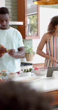 Dikey video: Kıvırcık saçlı genç bir kadın mutfakta tablet kullanıyor. Açık kahverengi bir teni, çizgili bir elbisesi var ve gülümseyerek yemek hazırlıyor.