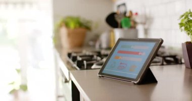 Bir tablet, akıllı bir ev uygulaması üzerinden ev enerjisi kullanımı hakkında renkli grafikler gösterir. Mutfak tezgahında duruyor. Etrafı saksı bitkileriyle ve yemek pişirme aletleriyle çevrili. Yavaş çekimde..