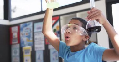 Okulda, sınıfta, genç bir Asyalı öğrenci kimyasal reaksiyonları inceliyor. Güvenlik gözlüğü takmış, biri yeşil, diğeri mor, değişmez bir sıvıyla iki beher tutuyor..