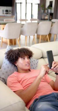 Dikey video: Genç bir çift ırklı adam elinde akıllı telefonuyla bir kanepede uzanıyor. Kıvırcık kahverengi saçları, sıcak bir gülümsemesi var ve günlük mercan tişörtü giyiyor..