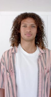 Dikey video: Neşeli bir gülümsemesi olan genç bir adam kendinden emin duruyor. Kıvırcık kahverengi saçlı, açık kahverengi tenli ve beyaz tişörtünün üzerine çizgili bir gömlek giyiyor.
