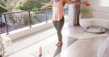 Yaşlı bir Afrikalı Amerikalı kadın evde yoga yapıyor. Kısa gri saçları, pembe bluzu ve gri taytı var. Duruşuna odaklanmış, değişmemiş, ağır çekimde.