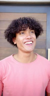 Dikey video: Genç bir çift ırklı adam neşeyle ışıldıyor. Kıvırcık saçları ve günlük pembe gömleği parlak ve pürüzsüz cildini tamamlıyor..