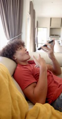 Çift ırklı genç bir adam kanepeye yaslanıyor, akıllı telefonuyla iletişim kuruyor. Kıvırcık saçları var, kırmızı gömlek giyiyor ve cihazıyla rahat bir ev ortamında meşgul görünüyor..