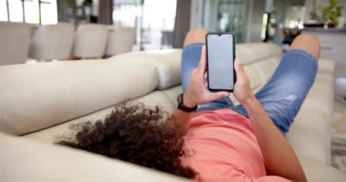 Genç bir melez kanepeye uzanmış, elinde akıllı bir telefon tutuyor. Kıvırcık siyah saçları var, pembe bir gömlek ve kot pantolon giyiyorlar, telefonlarıyla rahatlıyorlar, değiştirilmemişler.