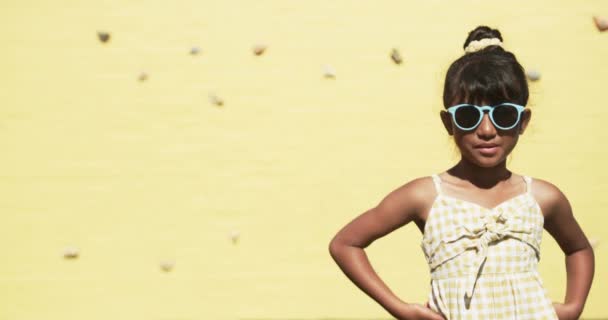 在学校里 一个年轻的学生站在一个黄色背景的复制空间前 她有一头乌黑的头发 结了个发髻 戴着蓝色的太阳镜 头戴无袖的格子上衣 毫发无损 — 图库视频影像