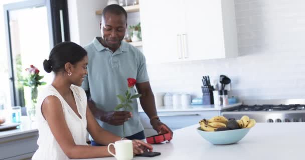 アフリカ系アメリカ人のカップルはキッチンで楽しい瞬間を共有し 贈り物を交換しています 彼は彼女に赤いバラをプレゼントします 両方とも 本物の笑顔で 変更されていない スローモーション — ストック動画