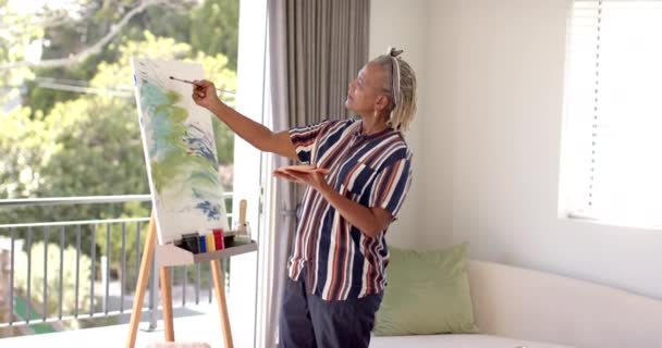 アフリカ系アメリカ人の女性が自宅のキャンバスに絵を描き 彼女の作品に焦点を当てた 彼女は灰色のドレッドロックを持っています ストライプのシャツを着用し 日光浴室に立っています 変更されていない ゆっくりとした動き — ストック動画