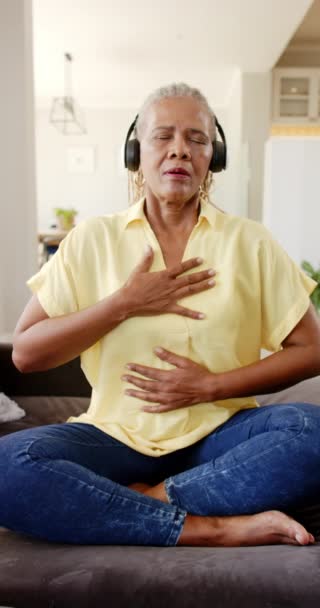 垂直ビデオ アフリカ系アメリカ人女性がヘッドフォンで音楽を楽しんでいます 彼女は座って 目を閉じて 胸に手をつけ 穏やかな家でリズムを感じている — ストック動画