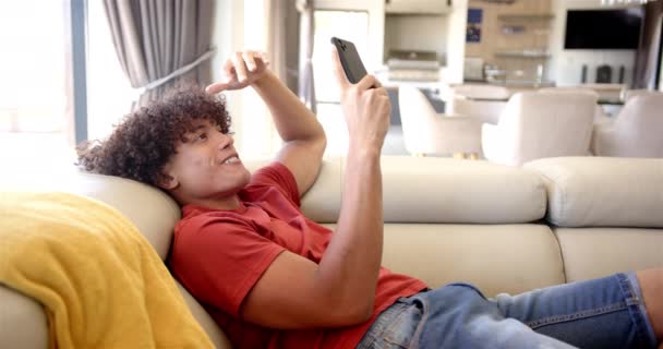 Çift Irklı Genç Bir Adam Evdeki Kanepede Uzanıyor Akıllı Telefonuyla — Stok video