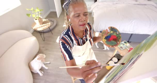 アフリカ系アメリカ人女性が自宅でキャンバスに絵を描いている 彼女は短い灰色の髪を持ち 暖かく微笑み エプロンでストライプのシャツを着ています 変更されていない ゆっくりとした動き — ストック動画