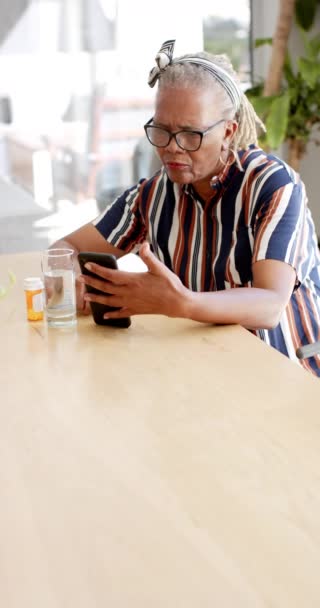 垂直录像 一名非洲裔美国妇女检查了她的智能手机 她的头发是灰的 戴着眼镜 穿着条纹衬衫 表情很专注 — 图库视频影像