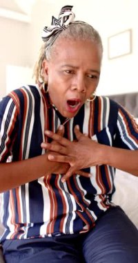 Dikey video: Yaşlı bir Afrikalı Amerikalı kadın göğüs ağrısı ya da kalp krizi geçiriyor. Kısa gri saçları var, çizgili bir gömlek giyiyor ve bir yatakta oturuyor..