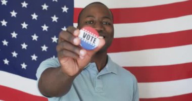 Genç bir Afrikalı Amerikalı seçmen rozeti gururla sergiliyor. Geniş bir gülümsemesi, kısa siyah saçları var ve açık mavi bir gömlek giyiyor, değişmemiş, ağır çekimde.