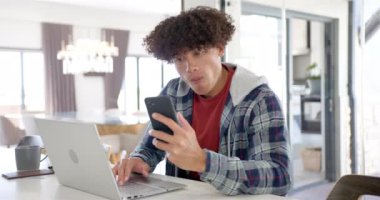 Çift ırklı genç bir adam akıllı telefonunu ve laptopunu evde kullanıyor. Kıvırcık saçları var, kırmızı bir kapüşonlu giyiyor. ve bilgisayarına odaklanıyor..