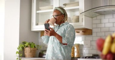 Yaşlı bir Afrikalı Amerikalı kadın kahvesini yudumluyor ve modern mutfağında akıllı telefonunu okuyor. Kısa gri saçları, gözlükleri var ve açık mavi bluzu var. Değiştirilmemiş, ağır çekimde..