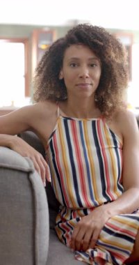 Dikey video: kıvırcık kahverengi saçlı melez bir kadın evinde kanepede sıcak bir şekilde gülümsüyor. Çizgili bir elbise giyiyor ve rahat bir şekilde kanepede oturuyor, rahat bir hava yayıyor..