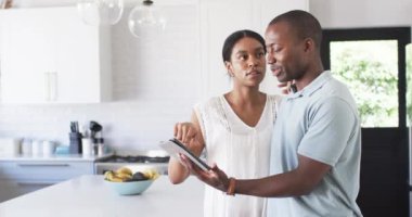 Çeşitli bir çift, mutfaklarında tablet kullanarak ve işaret ederek bir tartışmaya giriştiler. Afro-Amerikalı adam tableti tutarken düşünceli bir ifadeyle kadın, çerçevenin dışında bir şeye işaret ediyor..