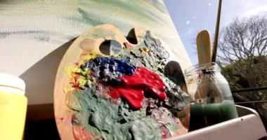 Bir palet ve resim güneş ışığında güneşlenir. Sanat malzemeleri düzgün dizilmiş, canlı renkler ve fırçalar arasında yaratıcı oturumu ima ediyor, değiştirilmemiş, yavaş çekim
