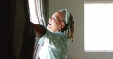 Kıdemli bir Afrikalı Amerikalı kadın evdeki perdeleri açıyor ve gün ışığını fotokopi odasıyla karşılıyor. Gri saçlı, neşeli bir yüzü var ve açık mavi bir gömlek giyiyor, değiştirilmemiş, ağır çekimde..