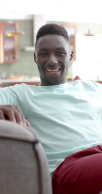 Dikey video: Genç bir Afrikalı Amerikalı adam sıcak bir şekilde gülümsüyor. Kısa siyah saçları, nane yeşili bir gömleği var ve evinde koltukta oturuyor..