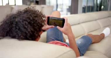 Çift ırklı genç bir adam bej bir kanepede uzanmış akıllı telefonuyla beyzbol seyrediyor. Akıllı telefona odaklanmış, bir oyun ya da video gösteriyor, değiştirilmemiş, yavaş çekim.