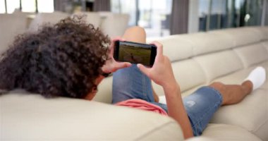 Çift ırklı genç bir adam bej bir kanepede uzanmış akıllı telefonuyla beyzbol seyrediyor. Akıllı telefona odaklanmış, bir oyun ya da video gösteriyor, değiştirilmemiş, yavaş çekim.