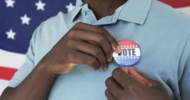 Afro-Amerikalı bir adam gömleğine seçmen rozeti takıyor. Parlak bir gülümsemesi var, açık mavi polo giyiyor, Amerikan bayrağına karşı, değiştirilmemiş, yavaş çekim