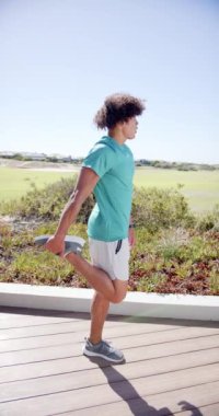 Dikey video: Çift ırklı bir adam ahşap bir patikada hamleler yapıyor. Açık bir gökyüzünün altında yeşil alanlarla çevrelenmiş halde, sağlık rutinine odaklanmış durumda..