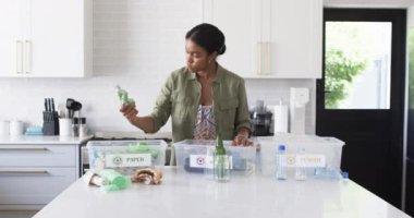 Genç bir Afrikalı Amerikalı kadın evdeki mutfakta geri dönüşümleri düzenliyor. Koyu renk saçları var, yeşil bir ceket giyiyor ve değiştirilmemiş, ağır çekimde bir şişe yerleştirmeye odaklanıyor..