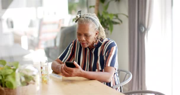 アフリカ系アメリカ人の女性は 自宅でスマートフォンに焦点を当て 薬を服用している 彼女は灰色の髪を編み ストライプのシャツを着て 明るいキッチンテーブルに座っています 変更されていない ゆっくりとした動き — ストック動画