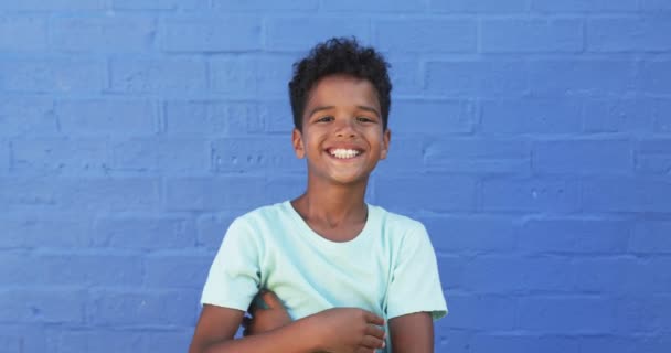 在学校里 一个年轻的非洲裔美国男孩在蓝色的背景下微笑 他有一头卷曲的黑头发 穿着一件浅蓝色的T恤衫 双手一动不动地摆姿势 — 图库视频影像