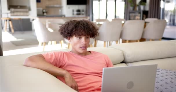 一个年轻的白人男子正在家里沙发上的笔记本电脑上工作 他有一头卷曲的棕色头发 穿着一件粉红色的T恤衫 一动不动地出现在银幕上 动作缓慢 — 图库视频影像