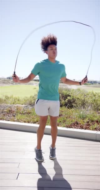 垂直ビデオ ロープをスキップする若い女性 青い空の背景 グリーンシャツ グレーのショートパンツ スニーカーを着て 彼のトレーニングに焦点を当てる — ストック動画