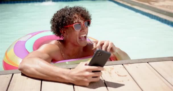 一个年轻的算命先生在家里带着智能手机的游泳池里放松一下 他一头卷曲的头发 戴着墨镜 一边喝着酒一边浏览着他的手机 一动不动的 慢吞吞的 — 图库视频影像