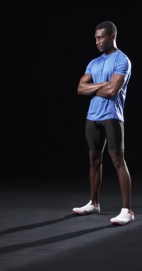 Dikey video: Afro-Amerikan erkek sporcu özgüvenle poz veriyor, siyah arka plan. Mavi gömlekli, siyah şortlu, güçlü, formda ve kollarını kavuşturmuş.