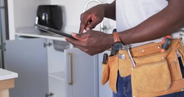 Afro-Amerikalı tamirci mutfakta tablet okuyor. Beyaz tişört giymiş, alet kemeriyle ekrana odaklanmış gibi görünüyor. Değiştirilmemiş, yavaş çekim..