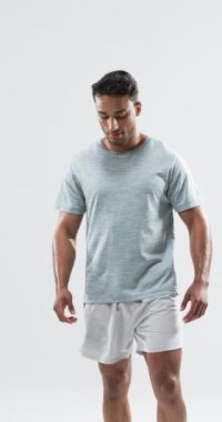 Dikey video: Çift ırklı erkek atlet pozu, beyaz arka plan. Açık gri tişört ve beyaz şort giyiyor, formda ve güçlü görünüyor.