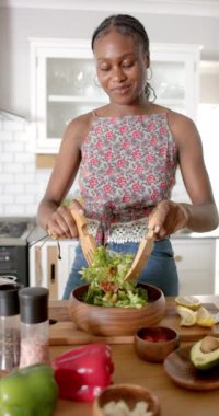 Dikey video: Afro-Amerikalı genç bir kadın mutfakta salata hazırlıyor. Saçları örgülü, çiçekli bluz giyiyor, yeşillikleri ahşap bir kaseye karıştırıyor.