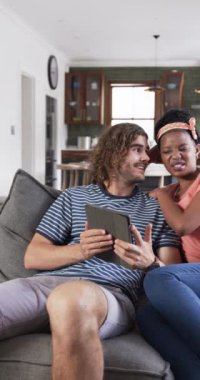 Dikey video: Evdeki kanepede oturup tablete bakan farklı bir çift. Saç bandı, kıvırcık saçlı Afrikalı Amerikalı kadın ve dalgalı saçlı beyaz adam.