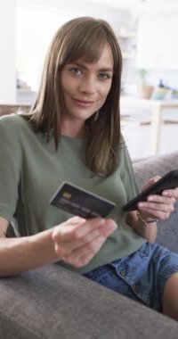 Dikey video: Kafkas kadının elinde kredi kartı var, evinde hafifçe gülümsüyor. Günlük kıyafetler giyiyor, omuz hizasında kahverengi saçlı ve açık renk makyajlı.