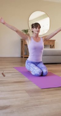 Dikey video: Kafkas kadın mor hasırda yoga yapıyor, aydınlatma odasında, evde. Orta yaşlı, kahverengi saçlı, açık renk bluzlu, desenli taytlı, kollarını kaldırmış.