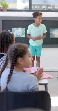 Dikey video: Okulda elinde tablet olan çocuk sunum yapıyor. Etrafını saran sınıf arkadaşları, arka planda sürdürülebilirlik posteriyle dikkatle dinliyorlar.