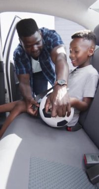 Dikey video: Afrikalı Amerikalı baba oğlunun emniyet kemerini takıyor, ikisi de gülümsüyor. Babasının kısa siyah saçları var, saat takıyor, oğlunun elinde futbol topu var, maç için heyecanlı.