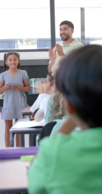 Dikey video: Okulda, genç kız sınıfına sunum yapıyor. Arkasında, sınıf arkadaşları dikkatle izlerken bir öğretmen alkış tutuyor.