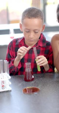 Okulda kırmızı kareli gömlek giyen çocuğun dikey videosu bir bilim deneyi yürütüyor. Odaklanmış, deney tüpünden bir şişeye sıvı boşaltıyor, masada kimya ekipmanları var..